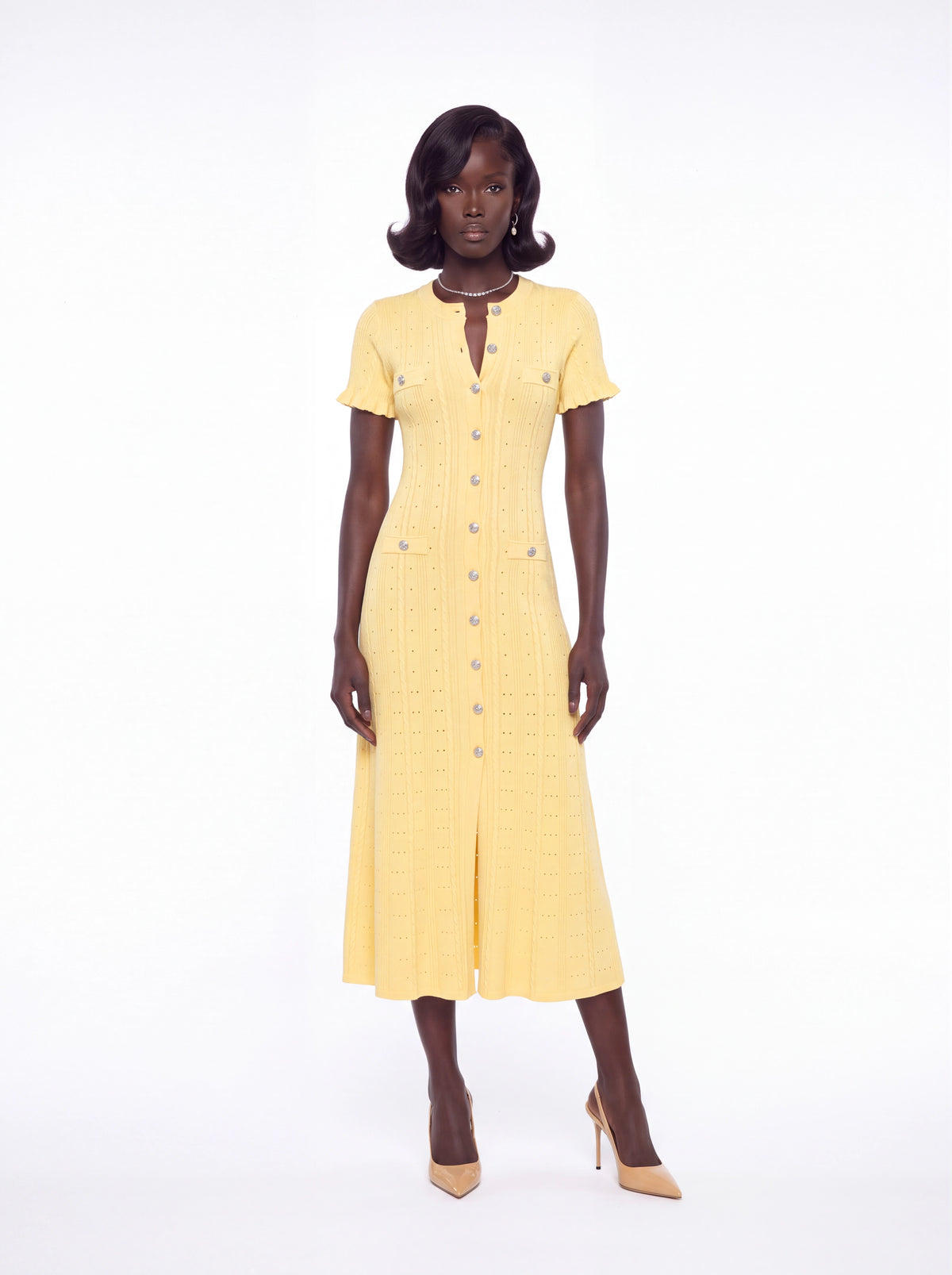 A woman modeling a pale yellow short-sleeve midi dress with a textured grid pattern, buttoned front, and split skirt, paired with beige heels against a white studio background.