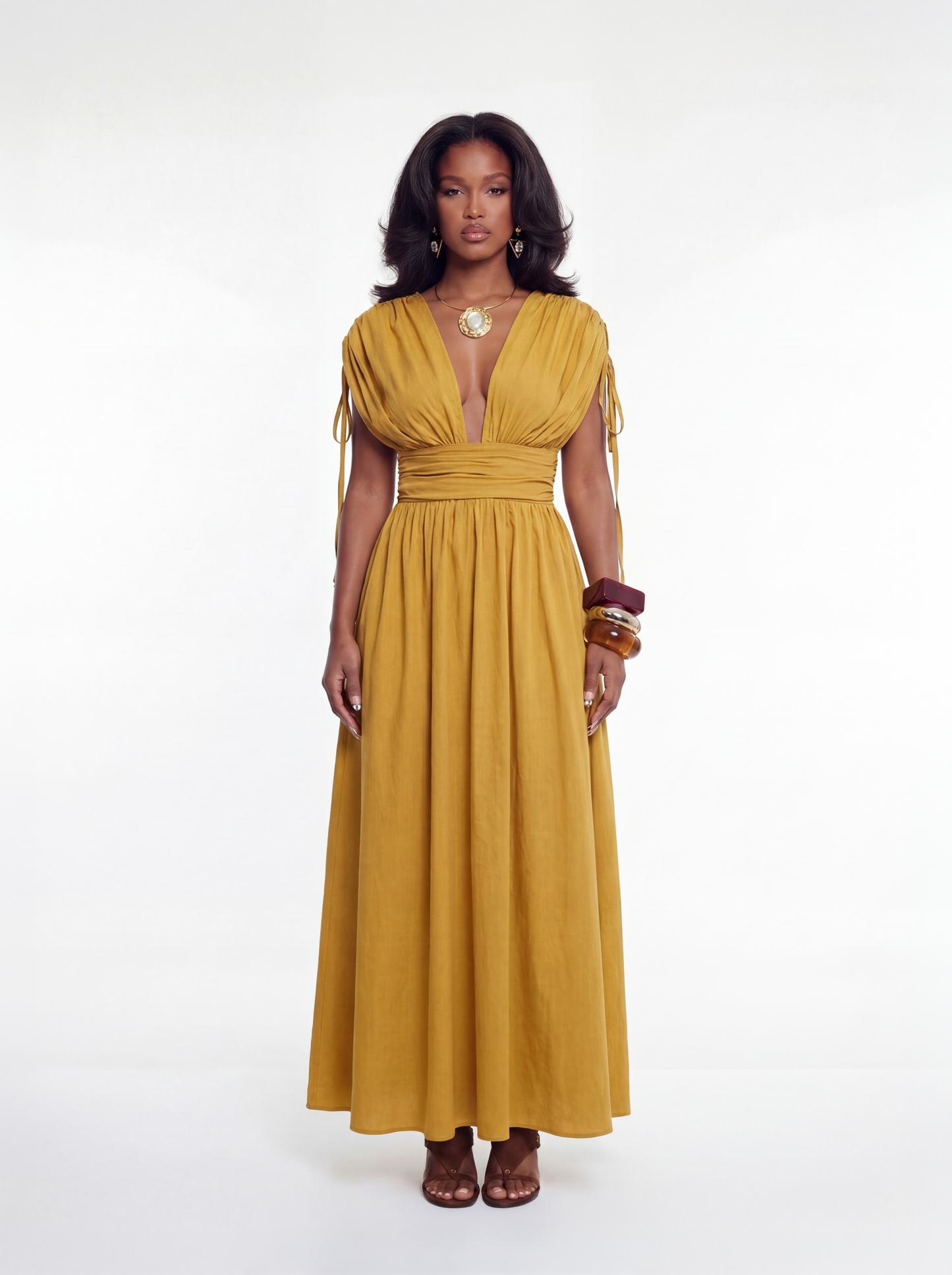 Woman wearing a mustard yellow maxi dress with a deep plunging neckline, gathered bodice, and cinched waist, featuring a flowing full-length skirt, styled with a statement necklace and stacked bangles.
