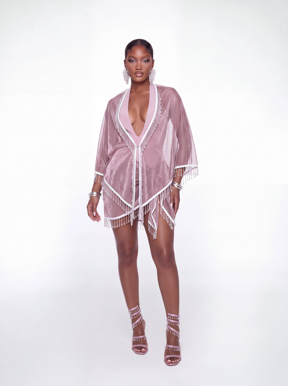 A model stands against a white studio background wearing a sheer dusty pink kimono cover-up with white fringe trim and matching pink strappy high-heeled sandals.