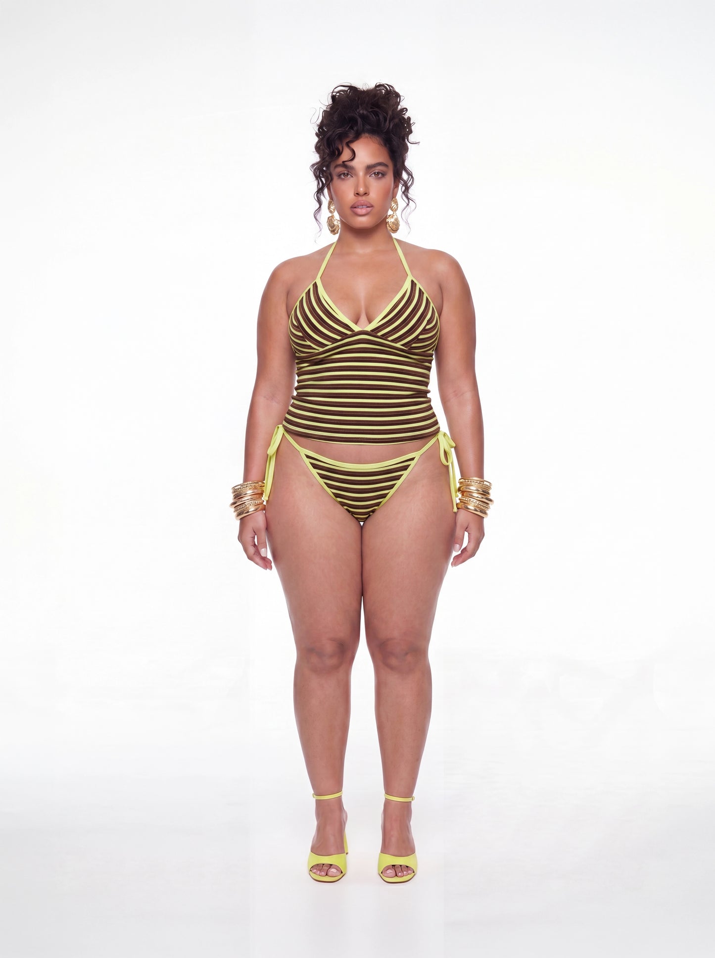 Curve model wearing a brown and yellow striped tankini swim set featuring a halter neckline, ruched bust detail, fitted tankini top, and matching side-tie bikini bottoms. Styled with gold statement earrings, stacked bangles, and yellow heels against a white studio background.