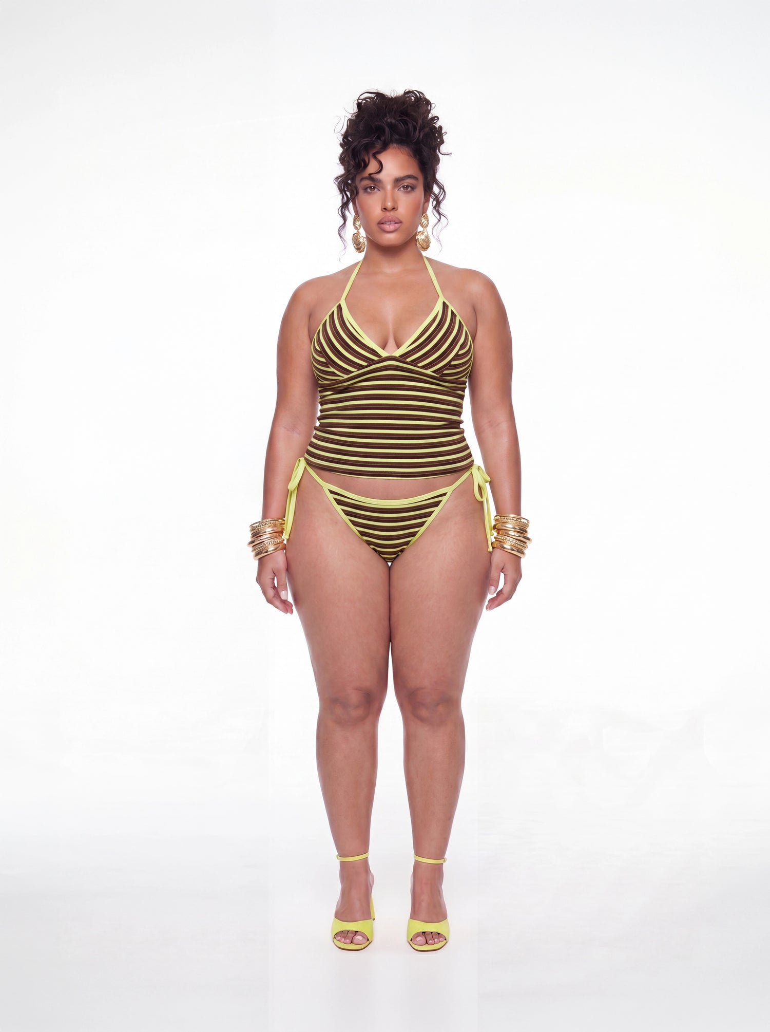 Curve model wearing a brown and yellow striped tankini swim set featuring a halter neckline, ruched bust detail, fitted tankini top, and matching side-tie bikini bottoms. Styled with gold statement earrings, stacked bangles, and yellow heels against a white studio background.