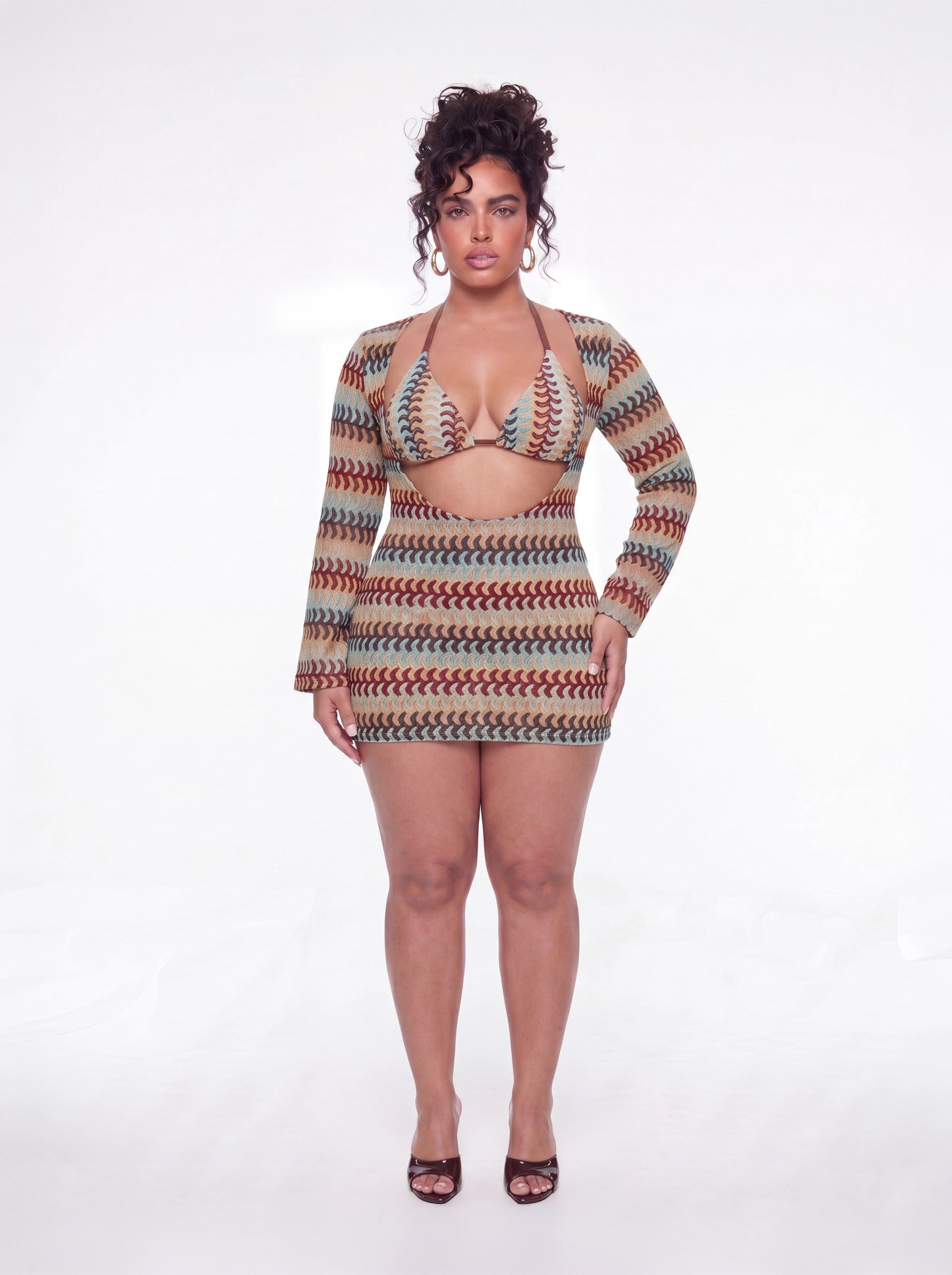 Curve model wearing a fitted long-sleeve mini dress featuring a multicolor wave print, triangle halter-style bodice, front waist cutout, and bodycon silhouette. Styled with gold hoop earrings and brown heels against a white studio background.