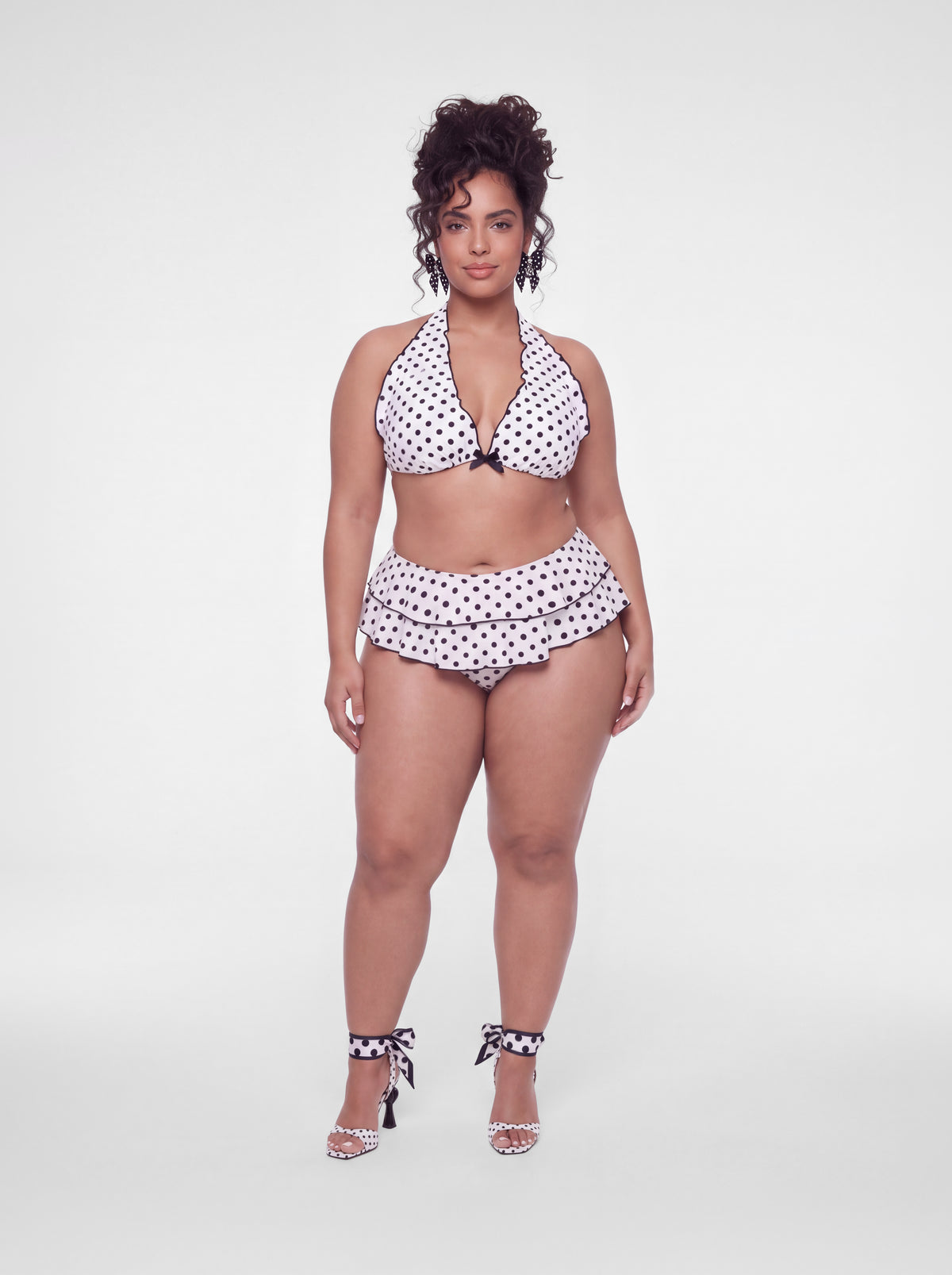 A woman modeling a white two-piece bikini with black polka dots, featuring a halter top and a ruffled high-waisted bottom, paired with matching polka-dot ankle-strap heels, against a light gray studio background.