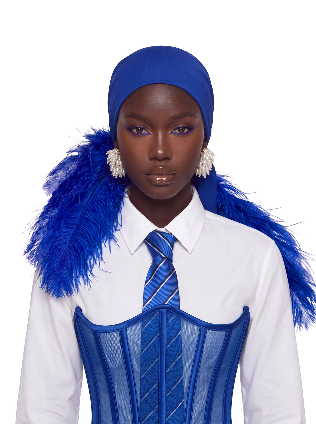 A model wearing a royal blue headscarf with attached blue feathers, a white collared shirt, a blue striped tie, and a blue sheer corset against a solid white background.