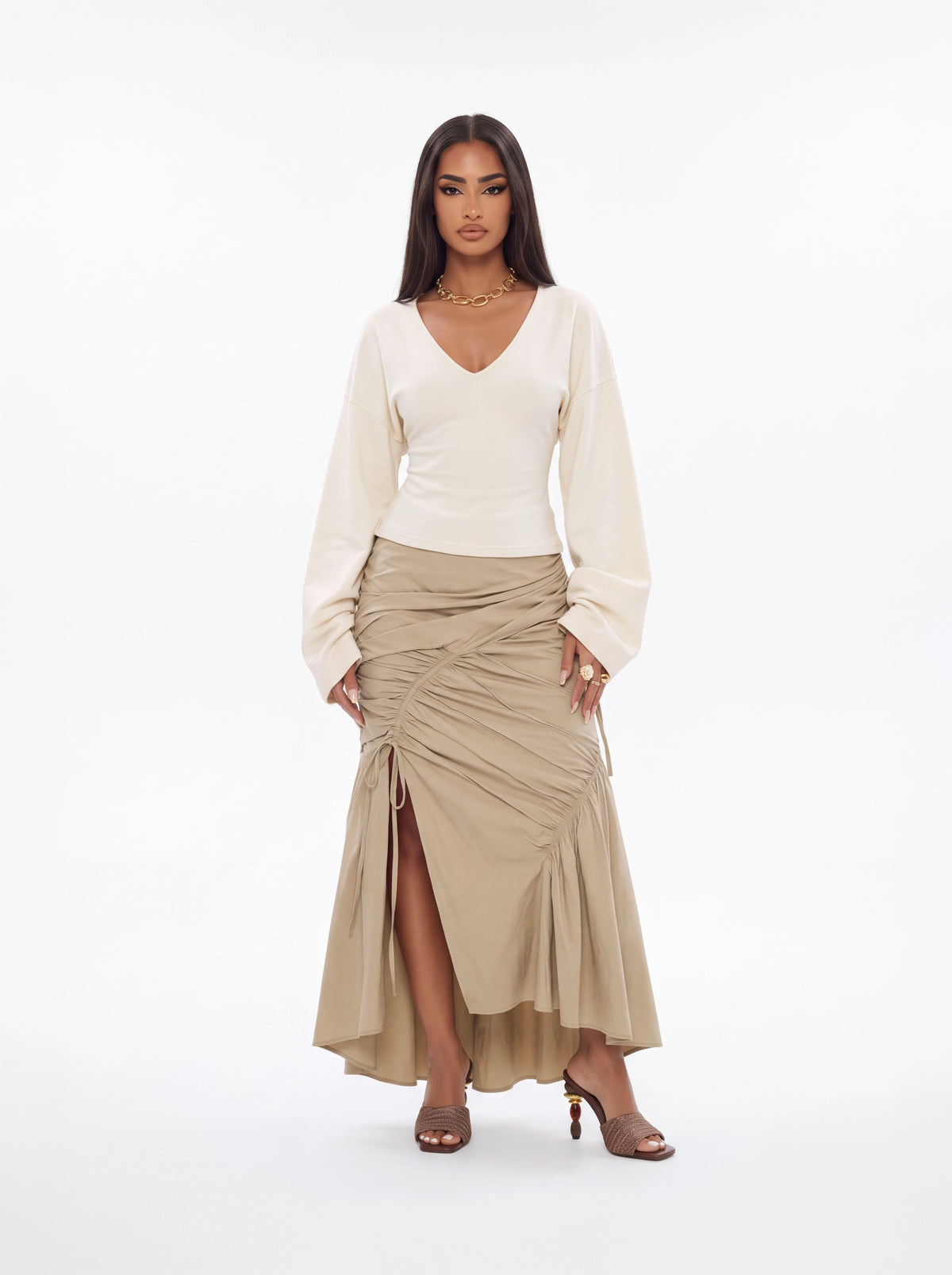 A model stands against a plain white studio background wearing a cream long-sleeve V-neck top and a beige ruched high-low skirt with a side slit.