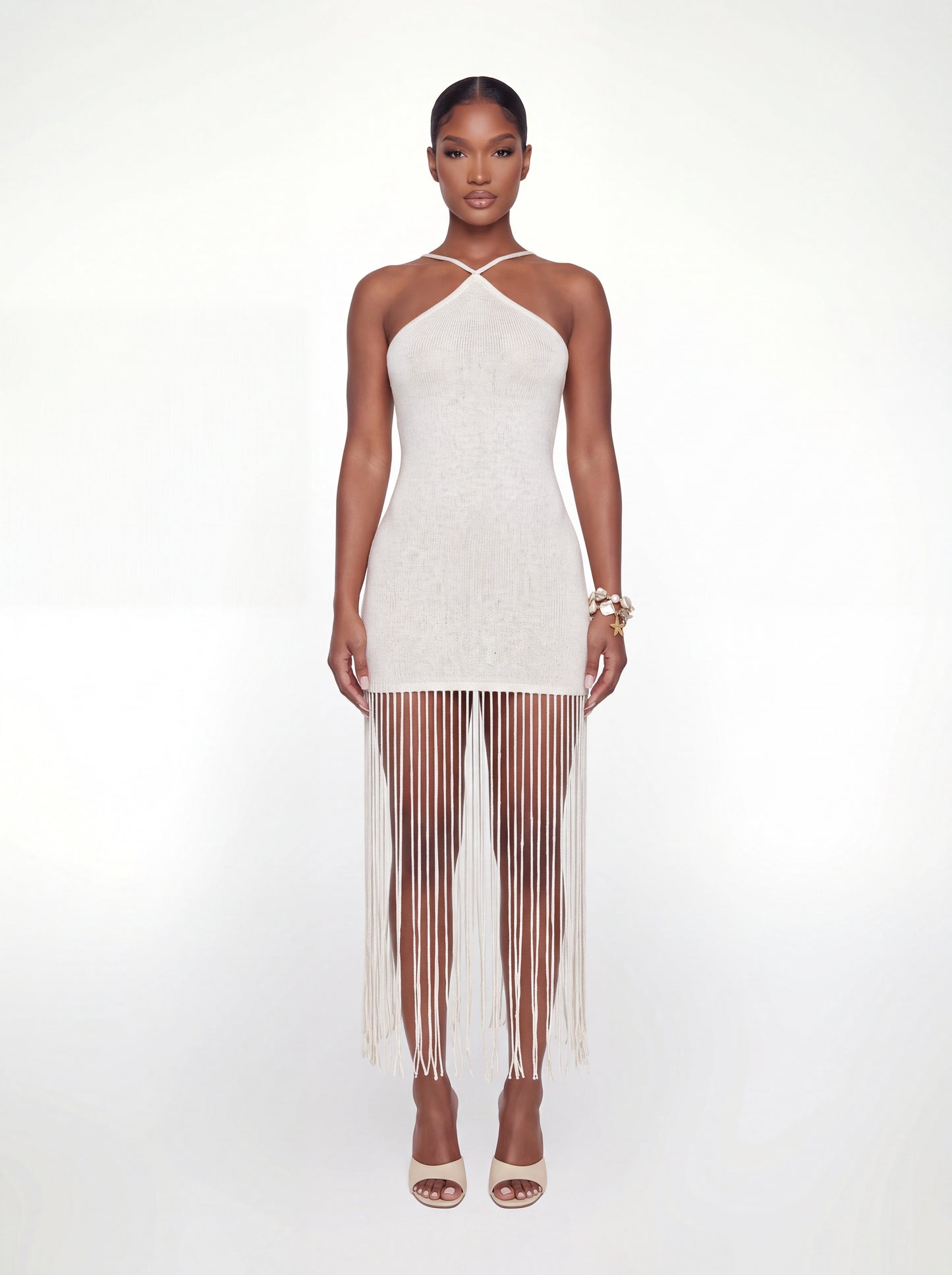 Front view of an ivory knit mini dress featuring a halter neckline with crossover straps, a fitted bodycon silhouette, and a dramatic long fringe hem. Styled with nude heeled sandals and shell bracelets against a white backdrop.