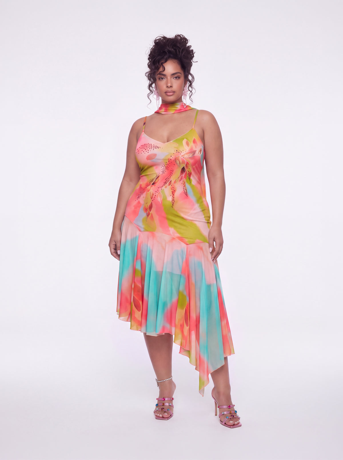 A woman modeling a colorful halter-neck dress with a tie-dye pattern in pink, yellow, green, and blue, paired with pink strappy high-heeled sandals against a plain white studio background.