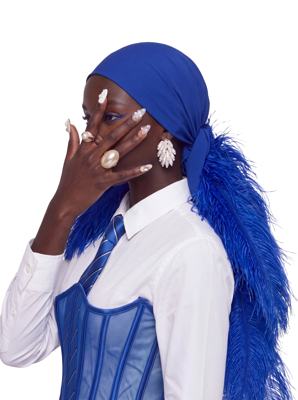 A woman wearing a royal blue feathered headscarf, a white collared shirt, a blue striped tie, and a blue corset vest, posed against a solid white background.
