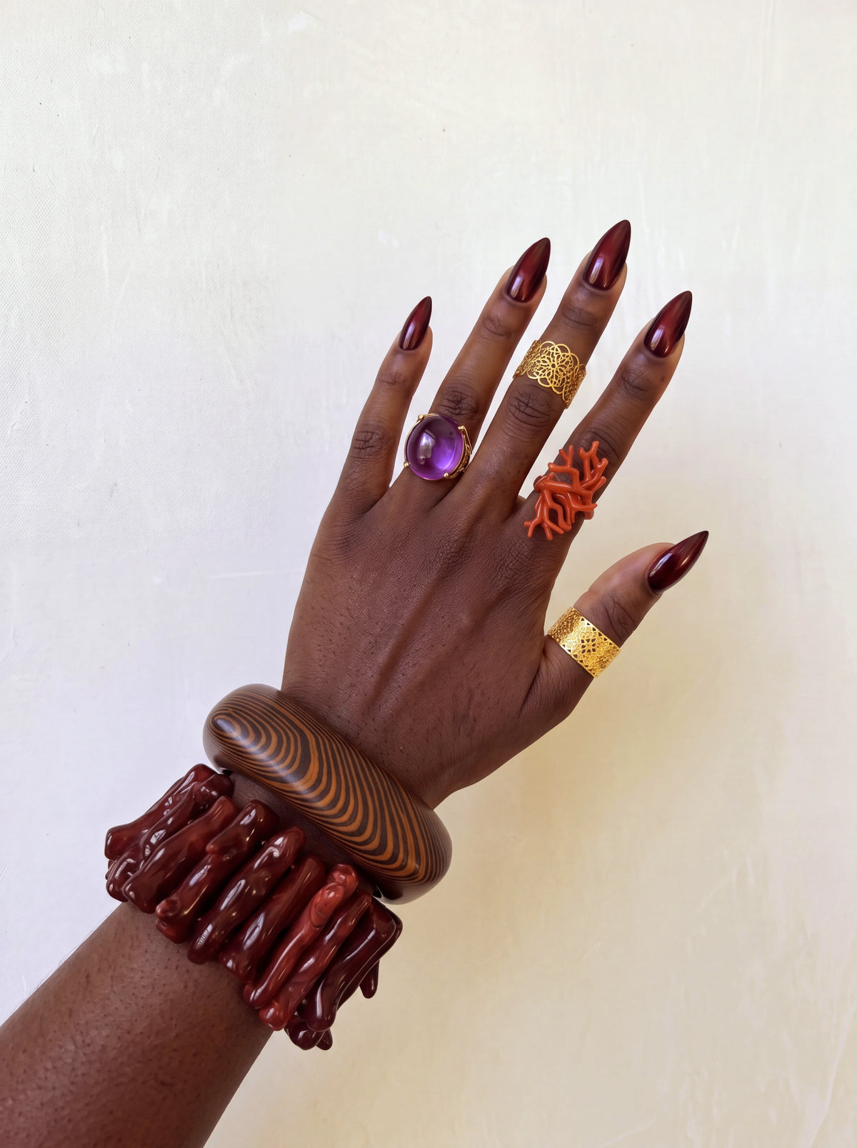 Close-up of a hand accessorized with a wood-grain bangle and chunky brown statement bracelet, layered with gold, purple gemstone, and orange coral-inspired rings and burgundy nails.