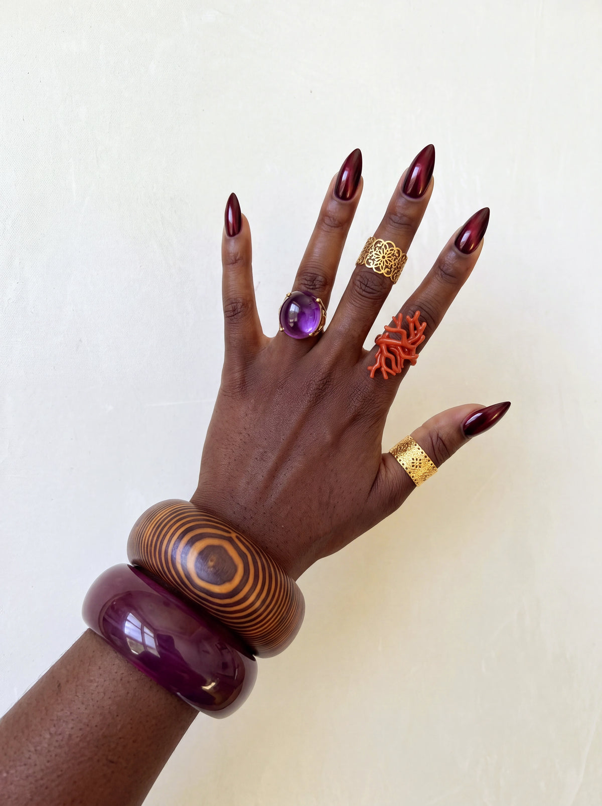 Close-up fashion shot featuring stacked oversized wood-grain bangles, colorful statement rings, metallic burgundy nails, and embellished strappy heels with beaded and tassel details, styled with a white mini dress.