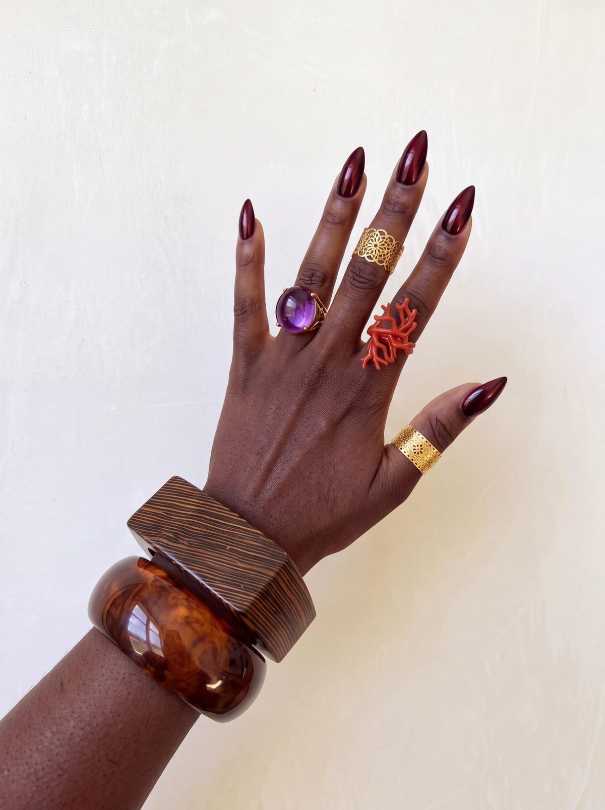 Hand styled with a rectangular wood-grain bracelet and glossy tortoiseshell bangle, paired with gold filigree, purple gemstone, and orange statement rings.