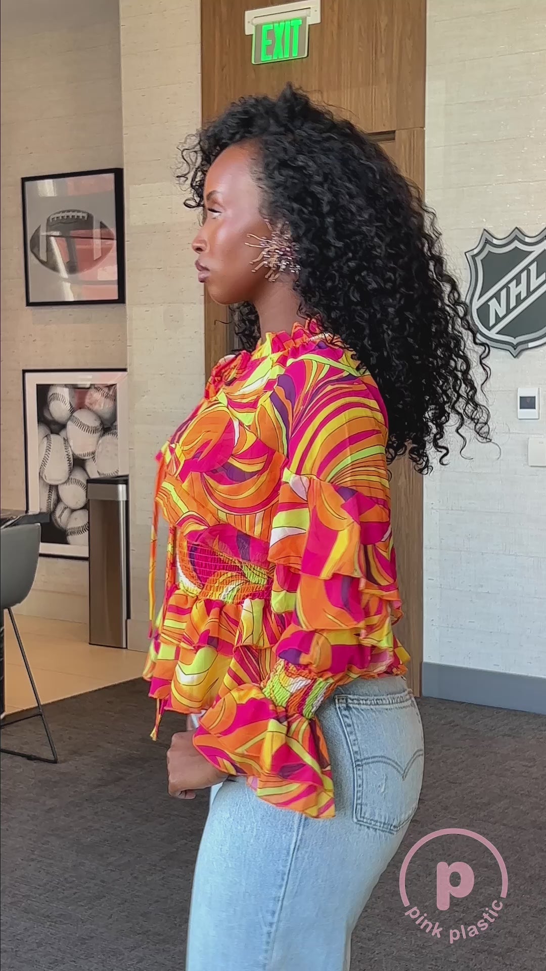 Model wearing the Vera Floral Top in coral, an orange and pink swirl print blouse with tiered ruffle sleeves, smocked waist and tie neck, styled with white denim pants. Showing multiple video cuts of model in the outfit in a room. 