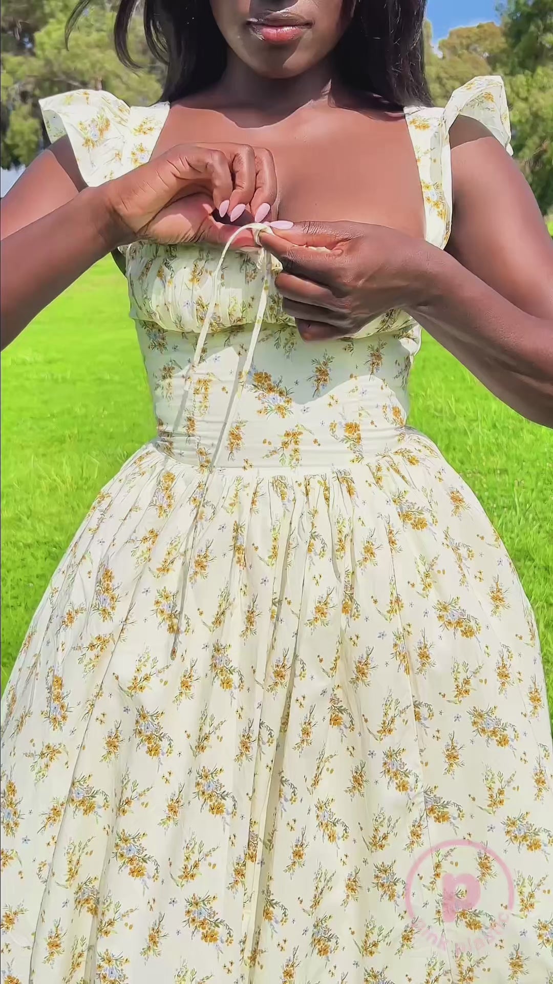 A woman models a cream dress with a delicate yellow floral print, ruffled shoulder straps, a fitted corset-style bodice, and a flowing skirt in a sunny park. She adjusts the neckline, lifts the skirt, and smiles while posing on the grass. Close-up shots highlight the dress’s details.