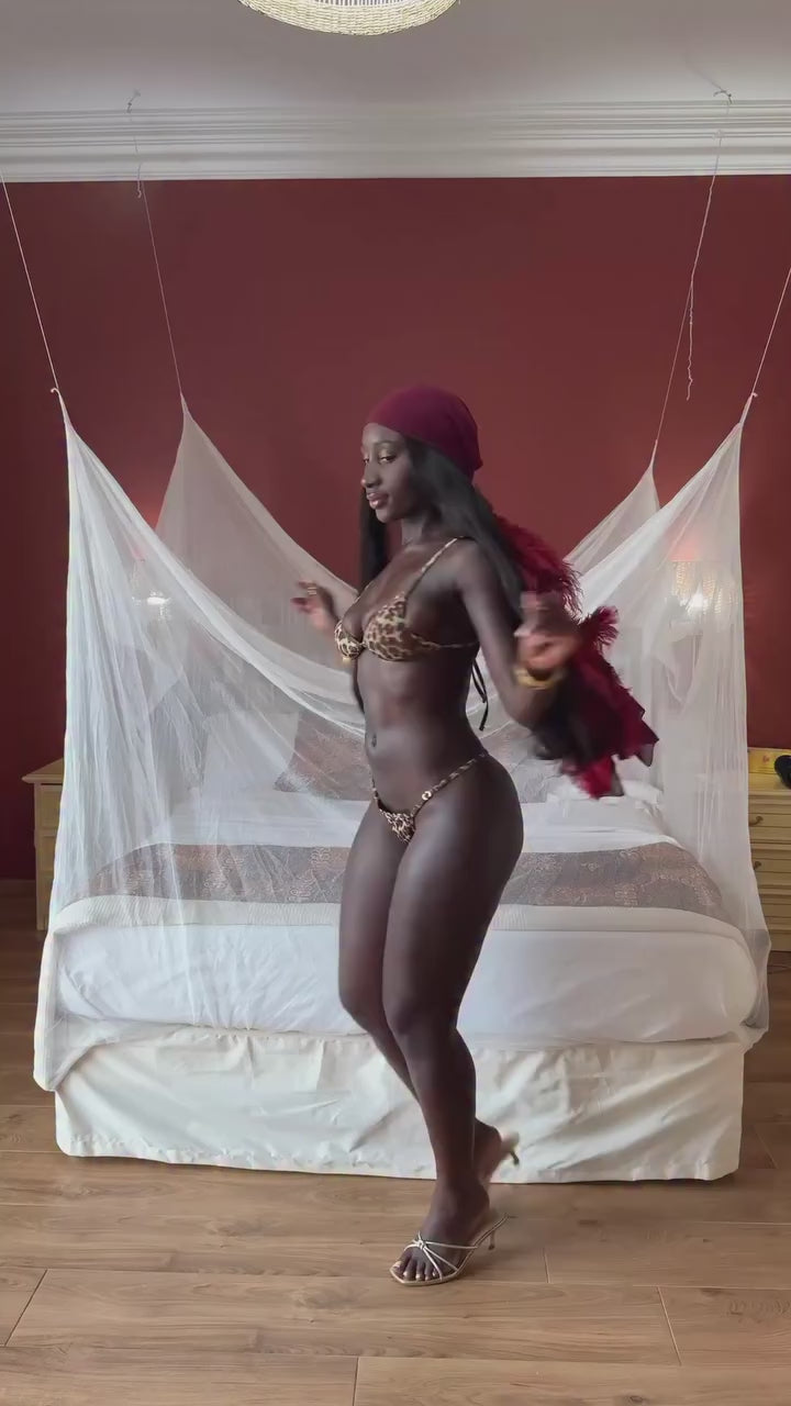 Model dancing and twirling in a two-piece animal-print swimsuit while wearing a burgundy headscarf trimmed with feathers.