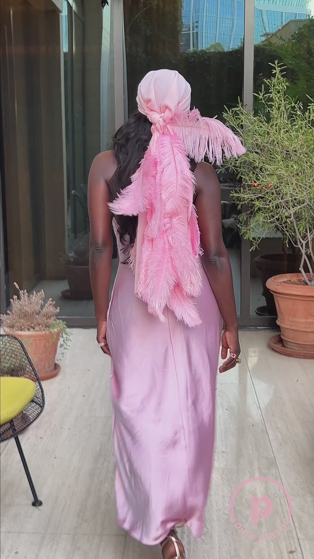 A woman models a pink satin slip dress with a matching feather-trimmed headscarf and green tassel earrings on a plant-filled patio. She poses standing, turns to show the scarf’s long feathered tails, and sits on a bench surrounded by greenery.