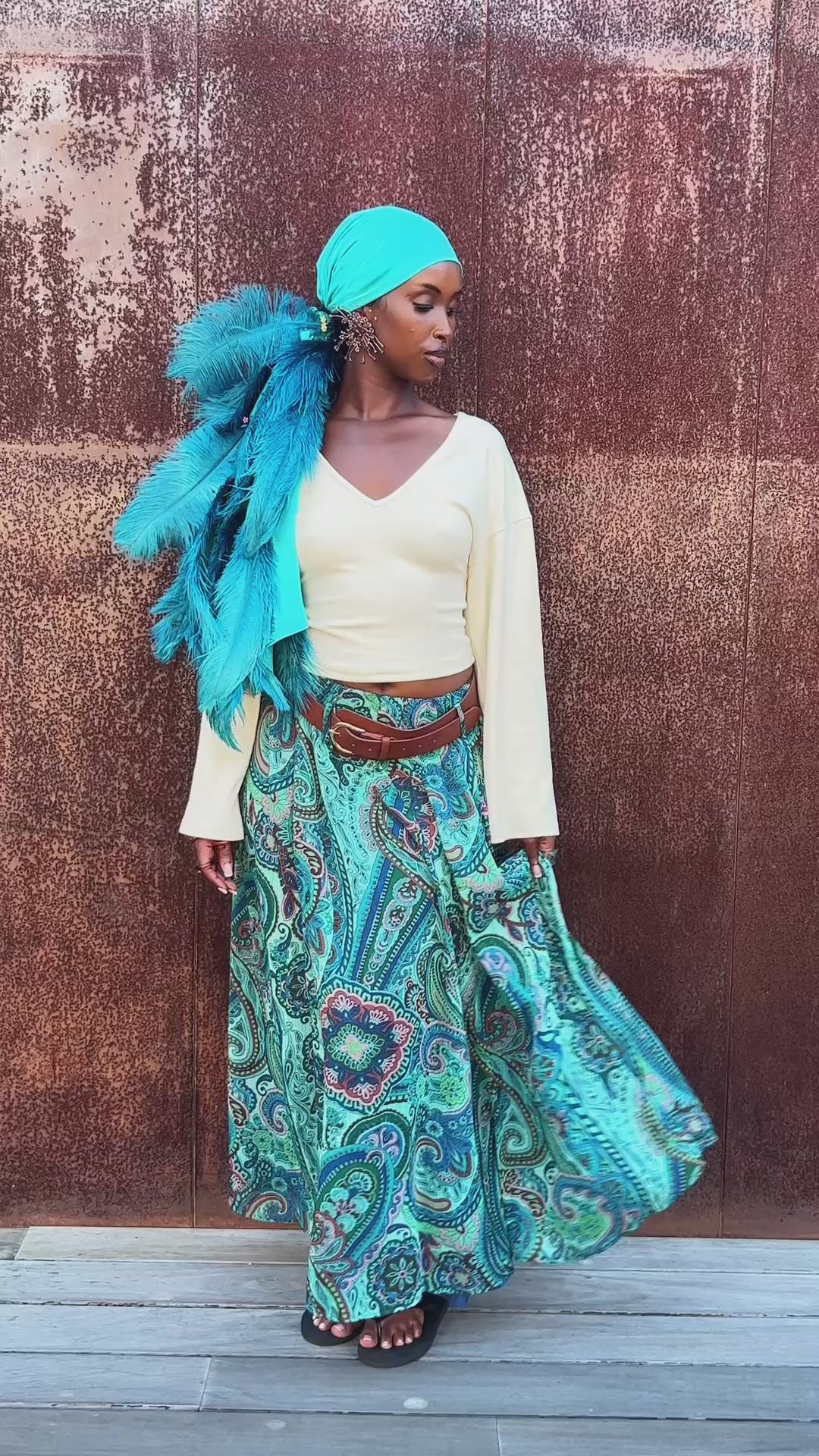 Model wearing a turquoise feather-trimmed headscarf, cream long-sleeve crop top, and flowing turquoise paisley maxi skirt, posing and moving the skirt in front of a textured brown wall.