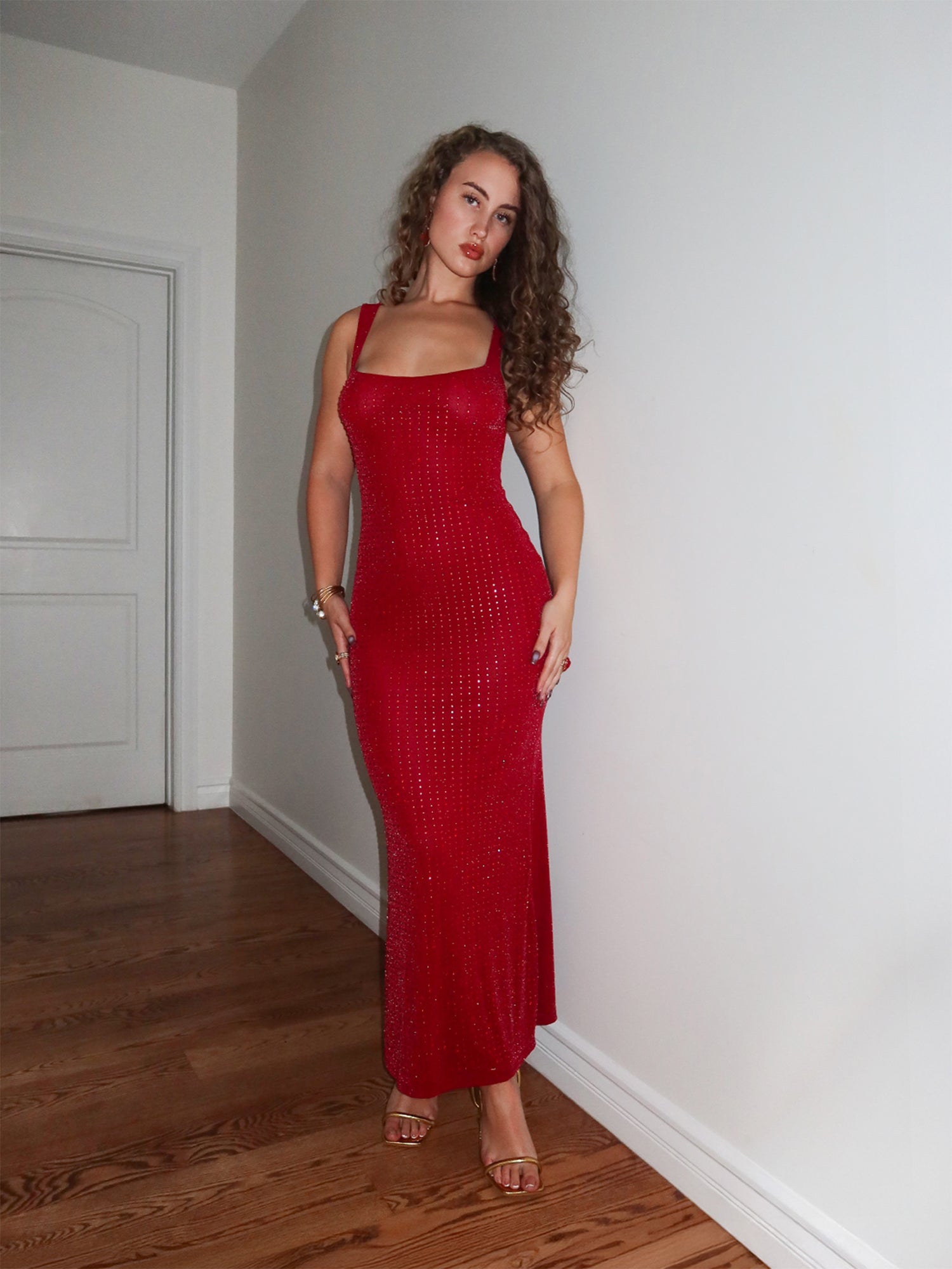 Fitted, floor-length red dress with a square neckline and wide shoulder straps. The dress features a low open back and a body-hugging silhouette. Shown on a model wearing gold heels and gold jewelry, photographed indoors against a white wall and staircase.