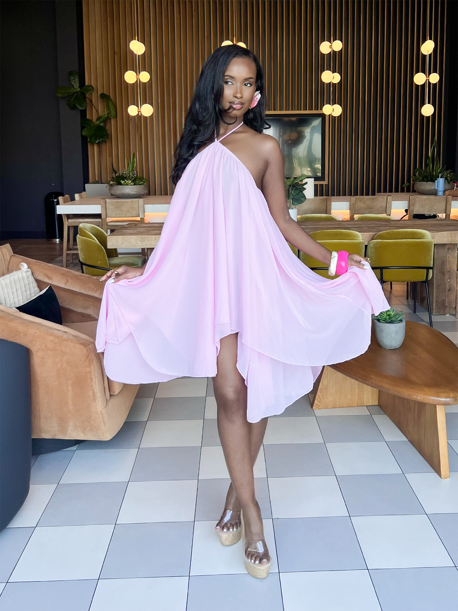 Woman posing indoors wearing a flowy pink halter mini dress with wedge heels in a modern lounge setting.