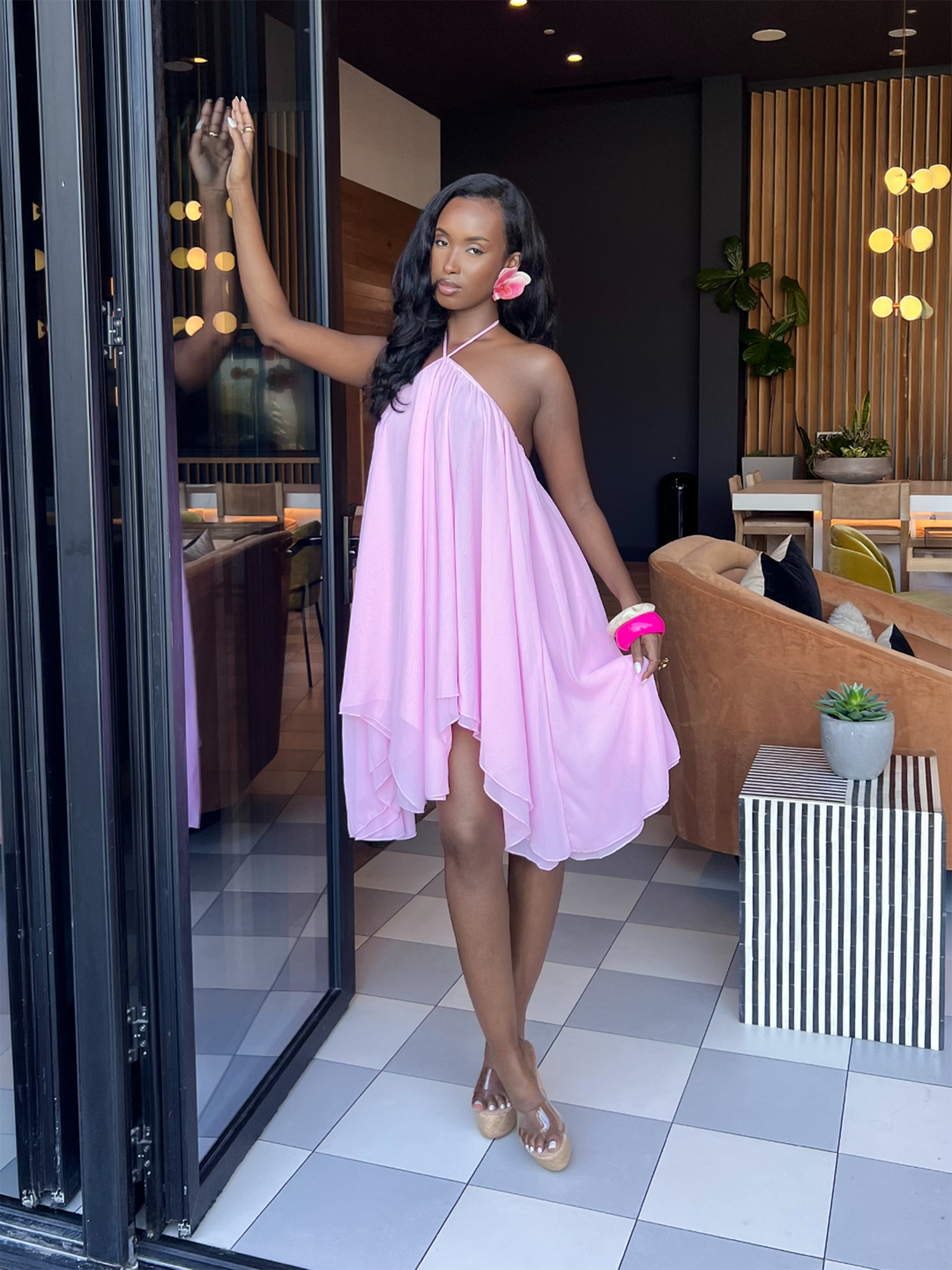 Woman standing in a doorway wearing a flowy pink halter dress with wedge heels in a modern lounge.