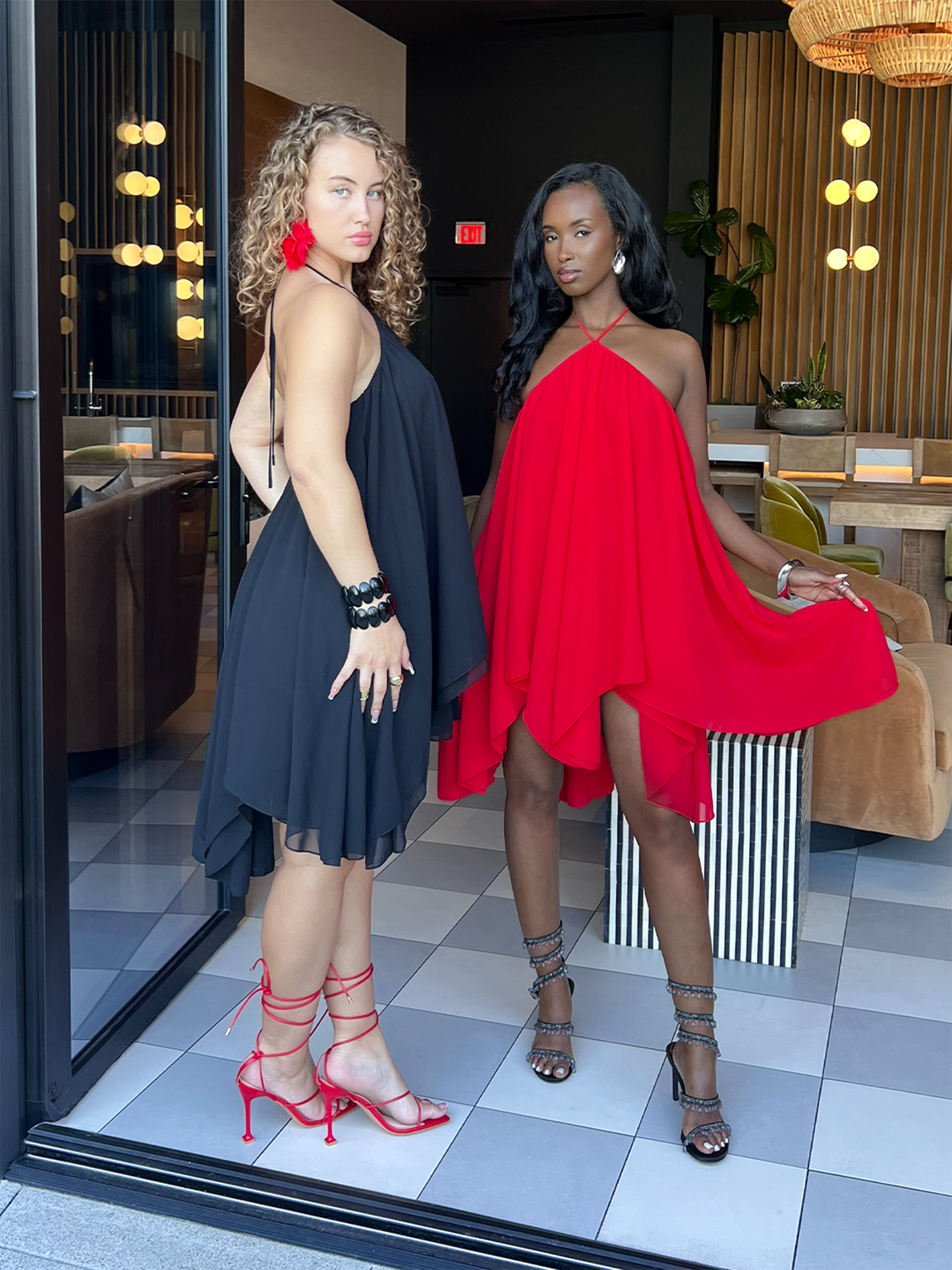 Two women posing in a modern lounge wearing flowy halter dresses—one in black and one in red—with strappy heels and statement accessories.
