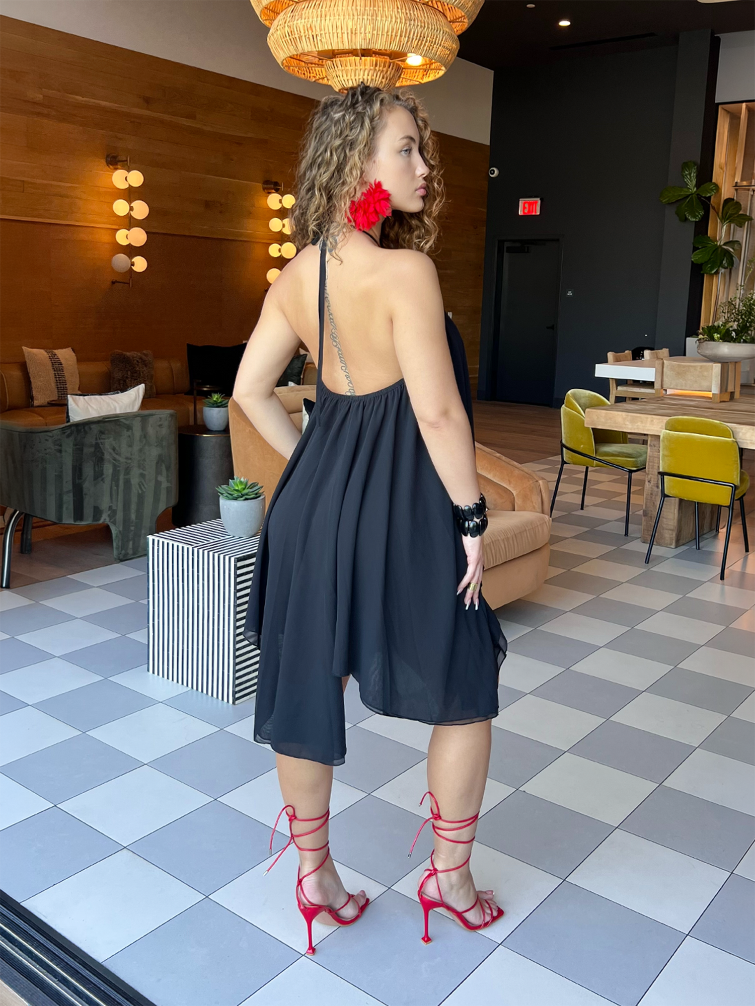 Woman posing from the back wearing a flowy black halter dress with an open back, styled with red strappy heels in a modern lounge.