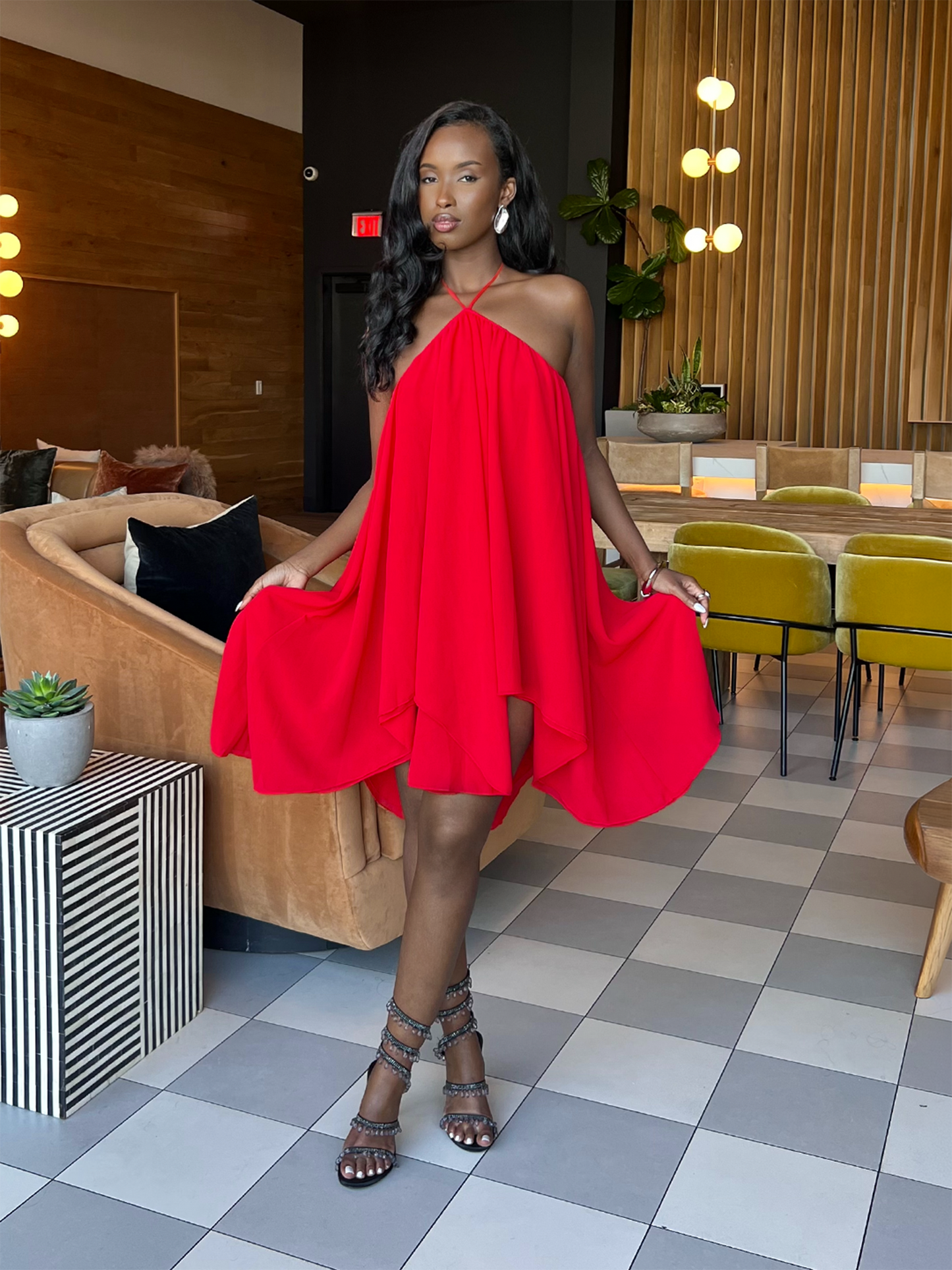 Model in a red dress standing in a modern indoor setting with checkered floor and furniture.