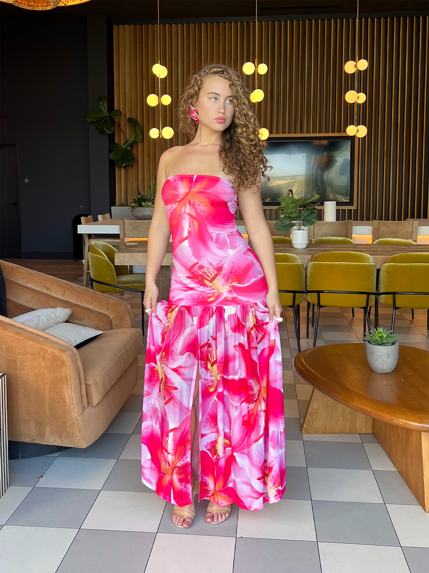 Model wearing a strapless pink floral maxi dress with a fitted bodice, drop waist, and flowy skirt with a front slit.